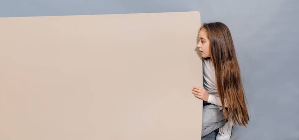 Large sheet of white cardboard. Little girl looks surprised at blank banner. Advertising. - Photo, Image