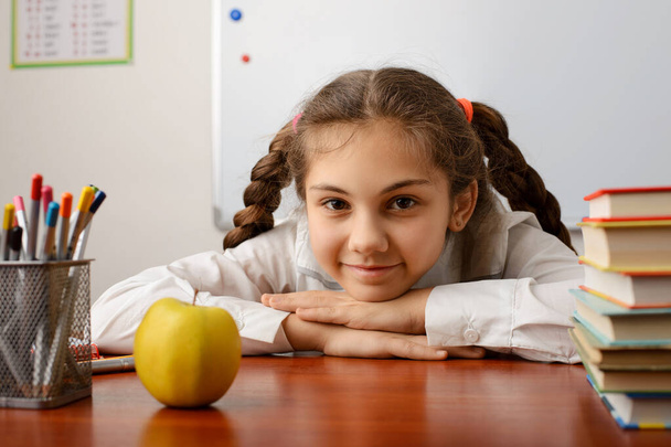 Juguetona colegiala alegre sentada cerca del escritorio con libros, lápices y una manzana durante la lección, mirando a la cámara - Foto, imagen