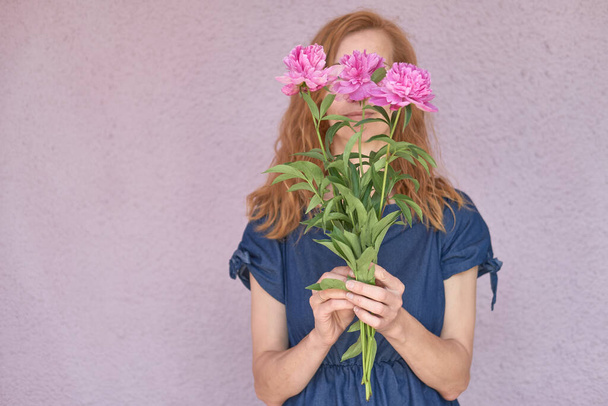 A faceless red-haired woman in a blue dress covers her face with pink peonies against a light wall. spring and summer concept. Copy space - Foto, immagini