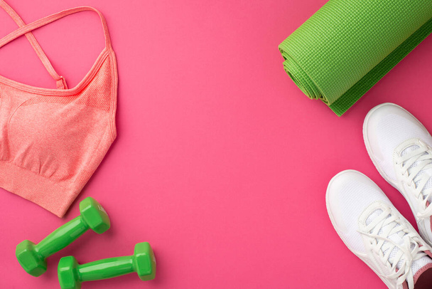 Концепция спортивных аксессуаров Фотография сверху зеленого коврика для упражнений розового спортивного топа и белых ботинок на изолированном розовом фоне с копирайтом - Фото, изображение