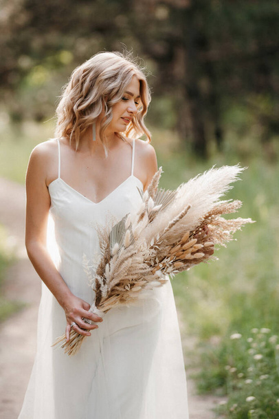 happy bride girl in a white light dress with a bouquet of dried flowers on a forest path - Photo, image