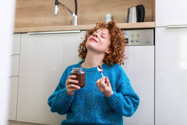 Eine junge Frau isst Schokolade aus einem Glas, während sie auf dem hölzernen Küchenboden sitzt. Niedliche Ingwermädchen frönen frechen Gesicht essen Schokoladenaufstrich aus Glas mit Löffel genießen jeden Schluck - Foto, Bild