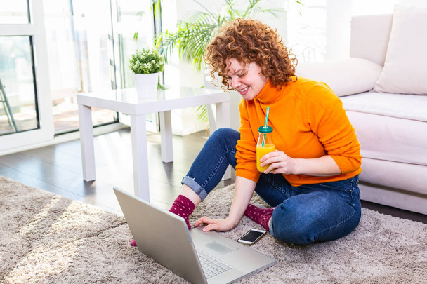 Képek egy fiatal vörös hajú nőről, aki narancslevet iszik és otthon szörfözik az interneten. Freelancer nő dolgozik laptop otthonról - Fotó, kép