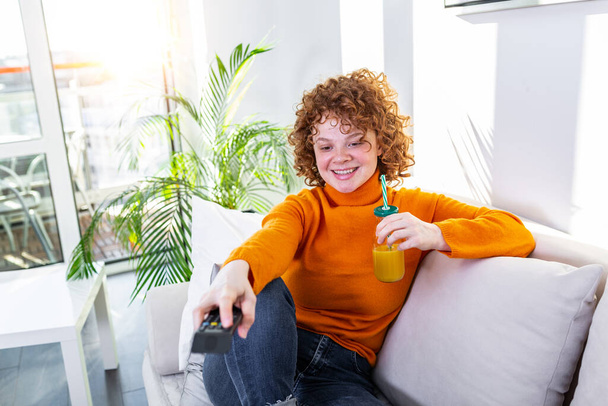Νεαρή γυναίκα με σγουρά κόκκινα μαλλιά κρατώντας τηλεχειριστήριο και βλέποντας τηλεόραση, αλλάζοντας κανάλια. Χαριτωμένο κορίτσι πίνει χυμό πορτοκαλιού και βλέπει τηλεόραση στο διαμέρισμά της - Φωτογραφία, εικόνα