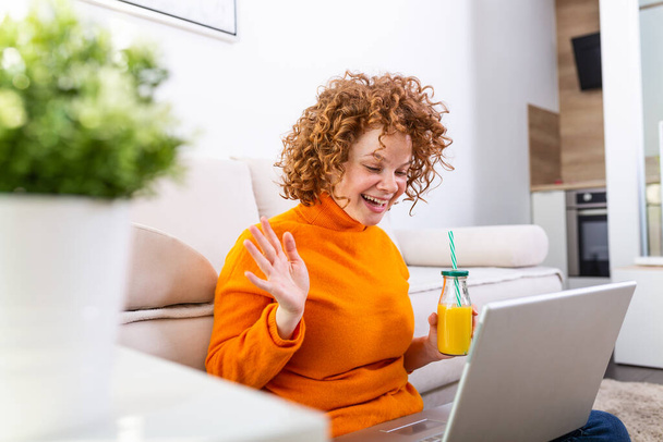 Młoda ruda rozmawiająca przez laptopa z rodziną. Młoda studentka za granicą rozmawiająca wideo z laptopa w domu i pijąca zdrowy sok pomarańczowy.. - Zdjęcie, obraz