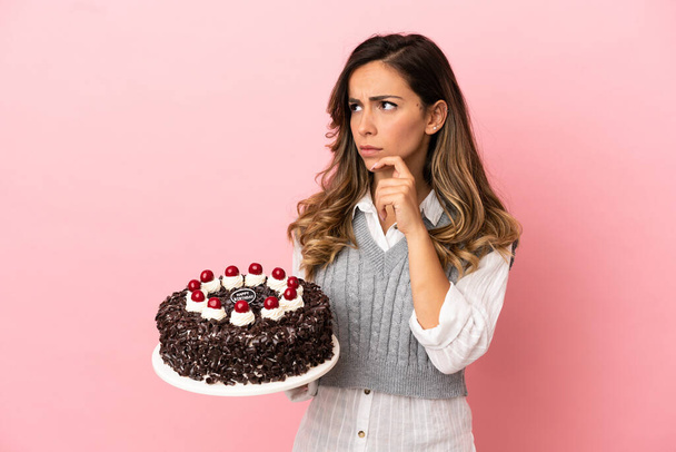 Giovane donna che tiene torta di compleanno su sfondo rosa isolato avendo dubbi e pensando - Foto, immagini