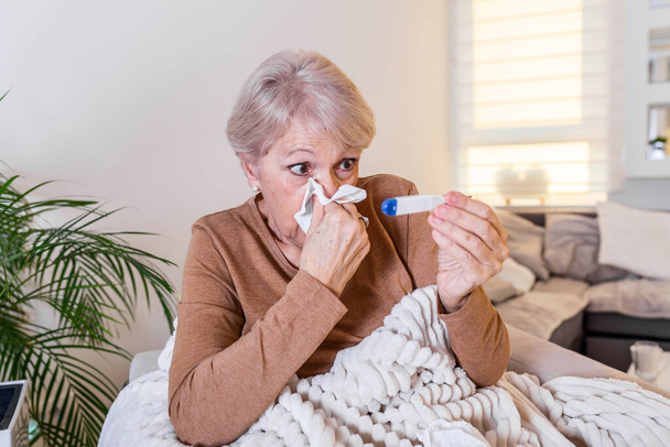 Sairaus, koronavirus ongelma käsite. Vanhempi nainen on sairas, kun flunssa makaa sohvalla katsomassa lämpömittarin lämpötilaa. COVID19-pandemia - Valokuva, kuva