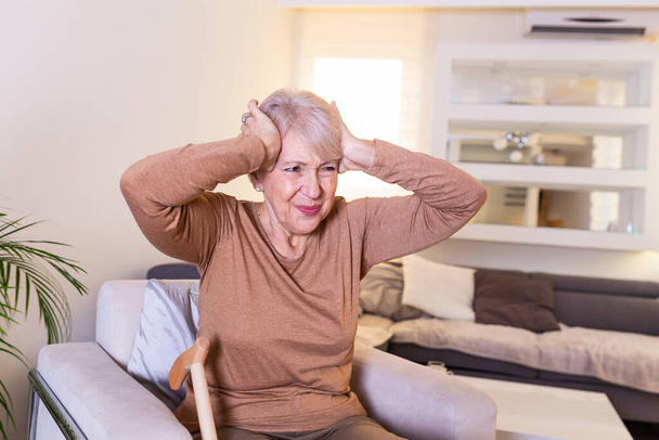 Starší žena sedící na bílé pohovce v domě dotýkající se rukou hlavy, zatímco bolest hlavy a pocit nevolnosti - Fotografie, Obrázek