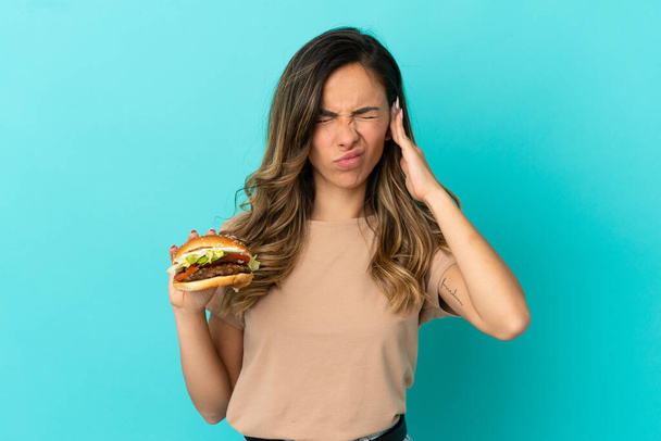 Young woman holding a burger over isolated background frustrated and covering ears - Foto, Bild