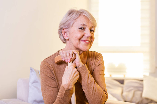 Portrait of beautiful senior woman with white hair and walking stick. Portrait of senior woman sitting on sofa at home. Smiling middle aged mature grey haired woman looking at camera - Photo, image