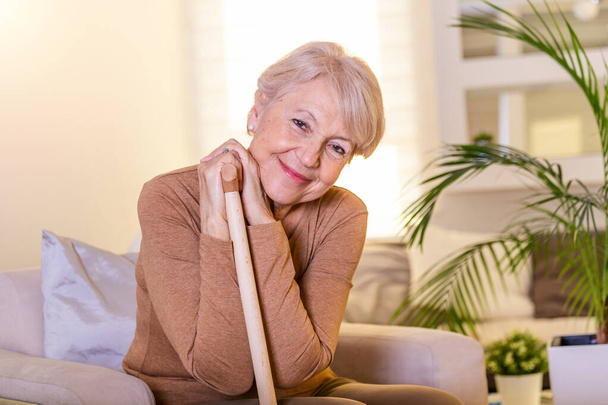 Ευχάριστη ηλικιωμένη γυναίκα που κάθεται με μπαστούνι. Συνταξιούχος γυναίκα με ξύλινο μπαστούνι στο σπίτι. Ευτυχισμένη ηλικιωμένη γυναίκα χαλαρώνει στο σπίτι κρατώντας μπαστούνι και κοιτάζοντας την κάμερα. - Φωτογραφία, εικόνα