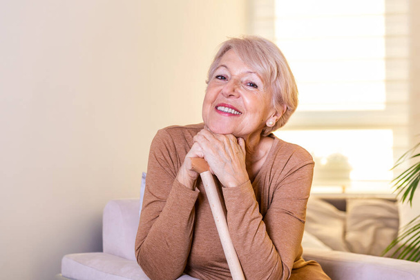 Usmívající se babička sedí na gauči. Portrét krásné usměvavé starší ženy s vycházkovou holí na světlém pozadí doma. Stará žena sedí s rukama na holi - Fotografie, Obrázek