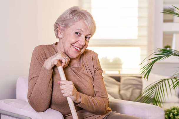 Porträt einer schönen Seniorin mit weißem Haar und Gehstock. Porträt einer Seniorin, die zu Hause auf dem Sofa sitzt. Lächeln mittleren Alters reifen grauhaarigen Frau Blick auf Kamera - Foto, Bild