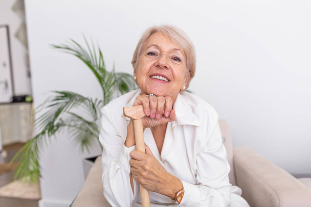 Portret van een mooie oudere vrouw met wit haar en wandelstok. Portret van een oudere vrouw die thuis op de bank zit. glimlachen middelbare leeftijd volwassen grijs harige vrouw kijken naar camera - Foto, afbeelding