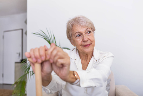 Elder lady sitting on the couch with wooden walking stick and smiling. Happy elderly woman relaxing on sofa and holding walking stick. Copy space. Senior woman looking thoughtful in a retirement home - Photo, Image