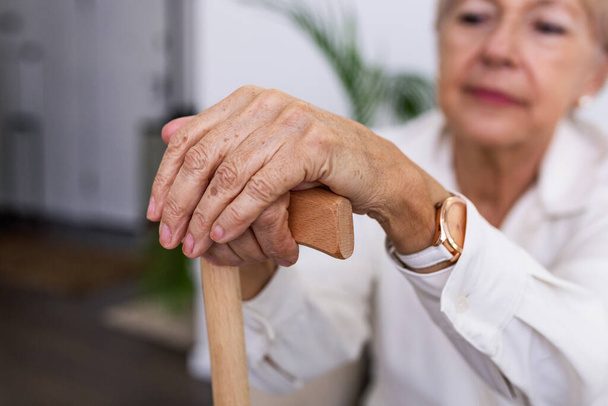 Hands of an old woman with a cane, Elder lady sitting on the couch with wooden walking stick. Cropped shot of a senior woman holding a cane in a retirement home - 写真・画像