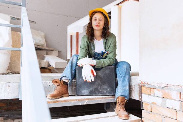 Having rest after hard working day. Live portrait of young beautiful woman, builder wearing helmet posing at construction site. Gender equality. Girl working at flat remodeling. Job, work, occupation - Fotó, kép