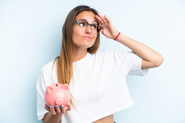 Young caucasian woman holding a piggybank isolated on blue background having doubts and with confuse face expression - Foto, imagen