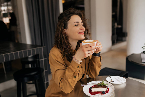Happy charming lady with curls wearing brown shirt enjoying morning coffee and dessert in cafe. Caucasian stylish lady is having breakfast in cafe.  - Фото, изображение