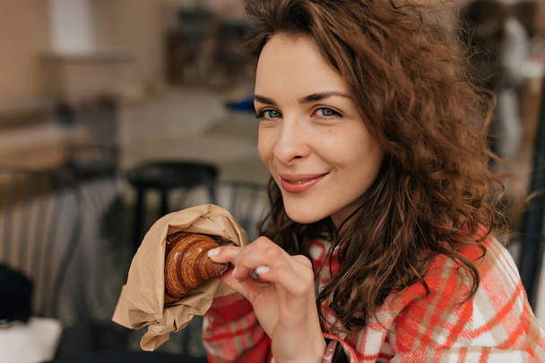 Close up profile portrait of curly elegant girl with perfect skin and big blue eyes with wavy hair wearing pink blouse eating croissant on summer terrace  - 写真・画像