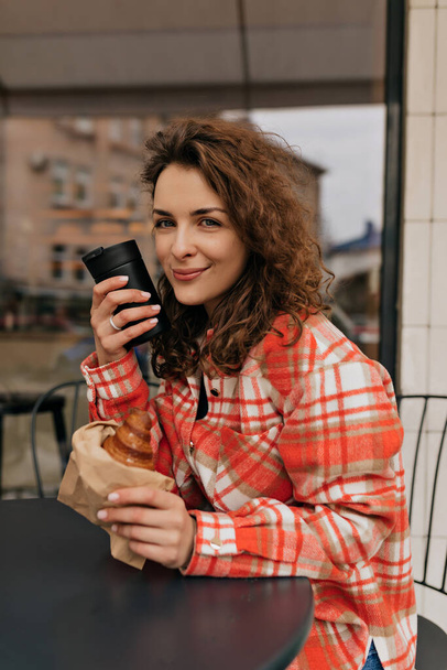 Πορτρέτο του σγουρά μελαχρινή καυκάσια γυναίκα σε πολύχρωμο φωτεινό πουκάμισο χαμόγελα ειλικρινά με καφέ στην καλοκαιρινή βεράντα και θέτει έξω. - Φωτογραφία, εικόνα