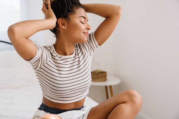 Young black woman smiling and holding her head while sitting on bed at home - Photo, image