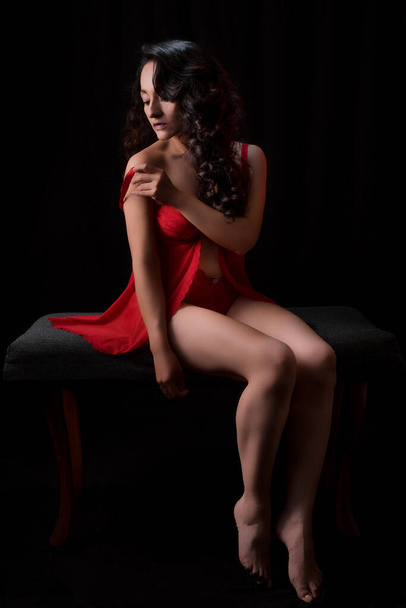 Όμορφη Λατίνα γυναίκα με κόκκινα εσώρουχα κάθεται σε ένα παγκάκι, με μαύρο φόντο, χαμηλή φωτογραφία κλειδί με φως τόπου είναι πολύ άνετη αναζητούν κομψό. - Φωτογραφία, εικόνα
