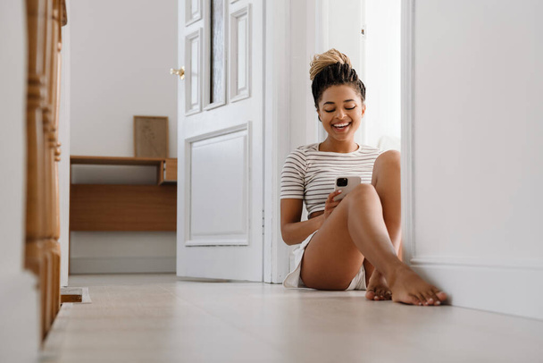 Young black woman using cellphone while sitting in doorway at home - Photo, image