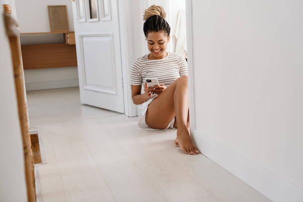Young black woman using cellphone while sitting in doorway at home - Zdjęcie, obraz