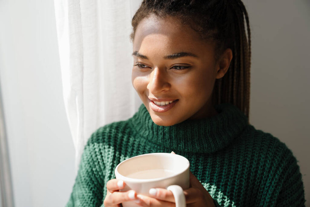 Young black woman with pigtails smiling while drinking tea at home - Φωτογραφία, εικόνα