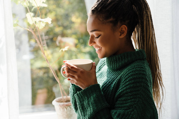 Young black woman with pigtails smiling while drinking tea at home - Foto, Imagen