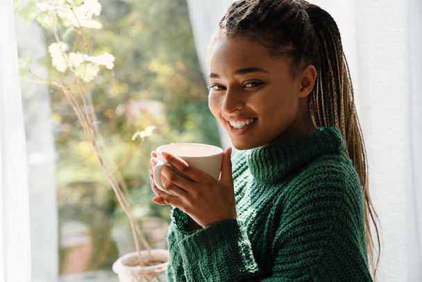 Young black woman with pigtails smiling while drinking tea at home - Photo, Image