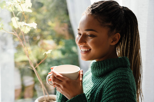 Young black woman with pigtails smiling while drinking tea at home - Foto, Bild