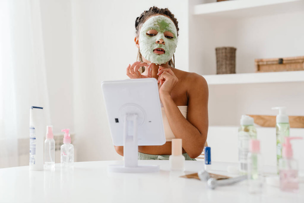 Young black woman with cosmetic mask looking in mirror at home - Fotó, kép