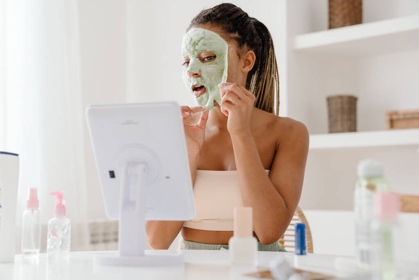 Young black woman with cosmetic mask looking in mirror at home - Foto, afbeelding