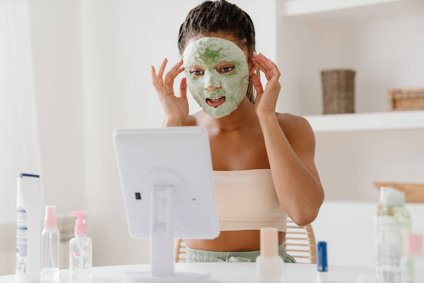 Young black woman with cosmetic mask looking in mirror at home - Фото, изображение