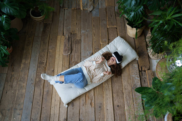 Young curious excited woman wearing 3d goggles resting on floor in greenhouse full of tropical houseplants, happy Italian female in VR headset lying on mattress enjoying virtual reality experience - Fotografie, Obrázek