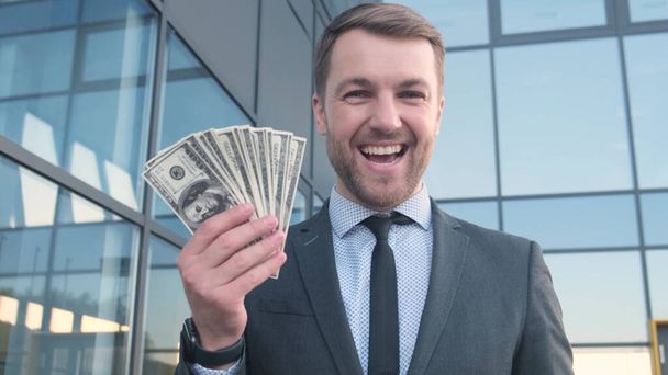 Πορτρέτο ενός χαμογελαστού επιτυχημένου άνδρα με κοστούμι που δείχνει με το δάχτυλό του ένα σωρό χαρτονομίσματα. - Φωτογραφία, εικόνα