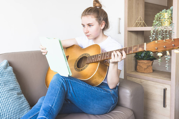 Vrouwelijke studente met akoestische gitaar en digitale tablet op een bank op gezellige thuisachtergrond. Jonge vrouw die online muzieklessen volgt, liedjes leert spelen of schrijft. Muziekhobby, muzikant, bank - Foto, afbeelding