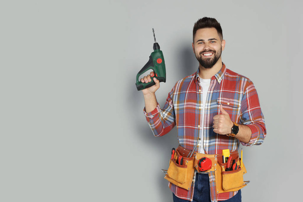 Young worker with power drill and tool belt on grey background. Space for text - Fotoğraf, Görsel