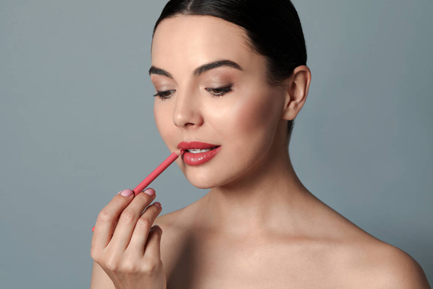 Pretty young woman applying beautiful pink lip pencil on grey background - Photo, image