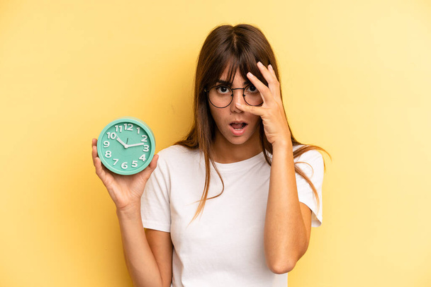hispanic woman looking shocked, scared or terrified, covering face with hand. alarm clock concept - Foto, Imagem