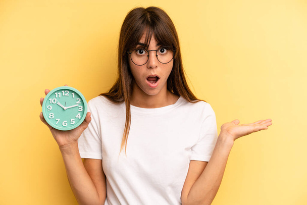 hispanic woman looking surprised and shocked, with jaw dropped holding an object. alarm clock concept - Фото, изображение