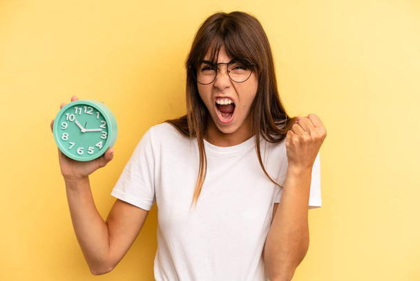 hispanic woman shouting aggressively with an angry expression. alarm clock concept - Фото, изображение