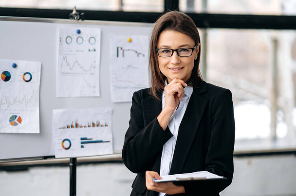 Portrait of confident elegant intelligent Caucasian business woman with glasses, in business clothes, entrepreneur, company ceo, stands in the office near the marker board, looks at the camera, smiles - Photo, image