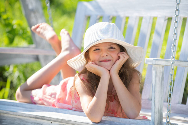 Little girl swinging on a summer sunny day. Child dreaming on swing. Child swinging on playground on sunny summer day in a park. Face of child girl in hat dreaming. Childhood, summer leisure - Photo, Image