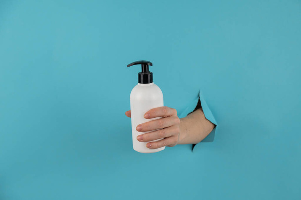 A womans hand sticks out of a hole in a blue paper background and holds a skin lotion pump - Foto, Bild
