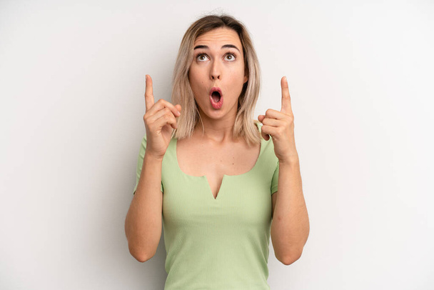 young adult blonde woman looking shocked, amazed and open mouthed, pointing upwards with both hands to copy space - Photo, image