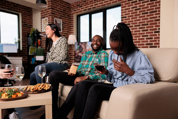 Cheerful diverse people laughing together at wine party in living room at home. Multiethnic group of friends smiling heartily while celebrating birthday event with alcoholic beverages and snacks. - Photo, Image