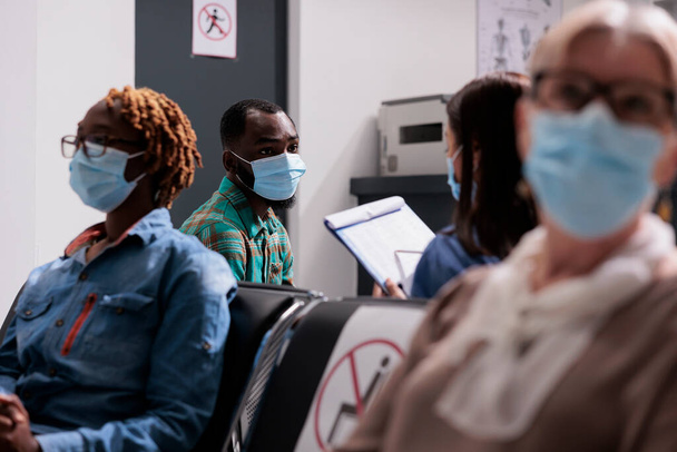 African american patient with face mask talking to nurse, sitting in waiting area at hospital reception lobby. Man doing healthcare consultation with medical worker at facility. - Foto, Imagem